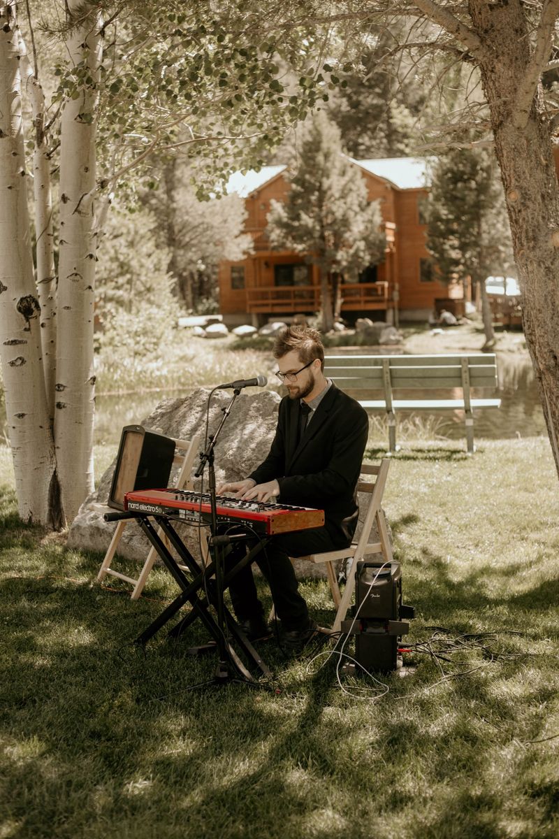 Max playing a red Nord keyboard and singing under aspen trees at a lakeside ceremony, a log cabin behind him