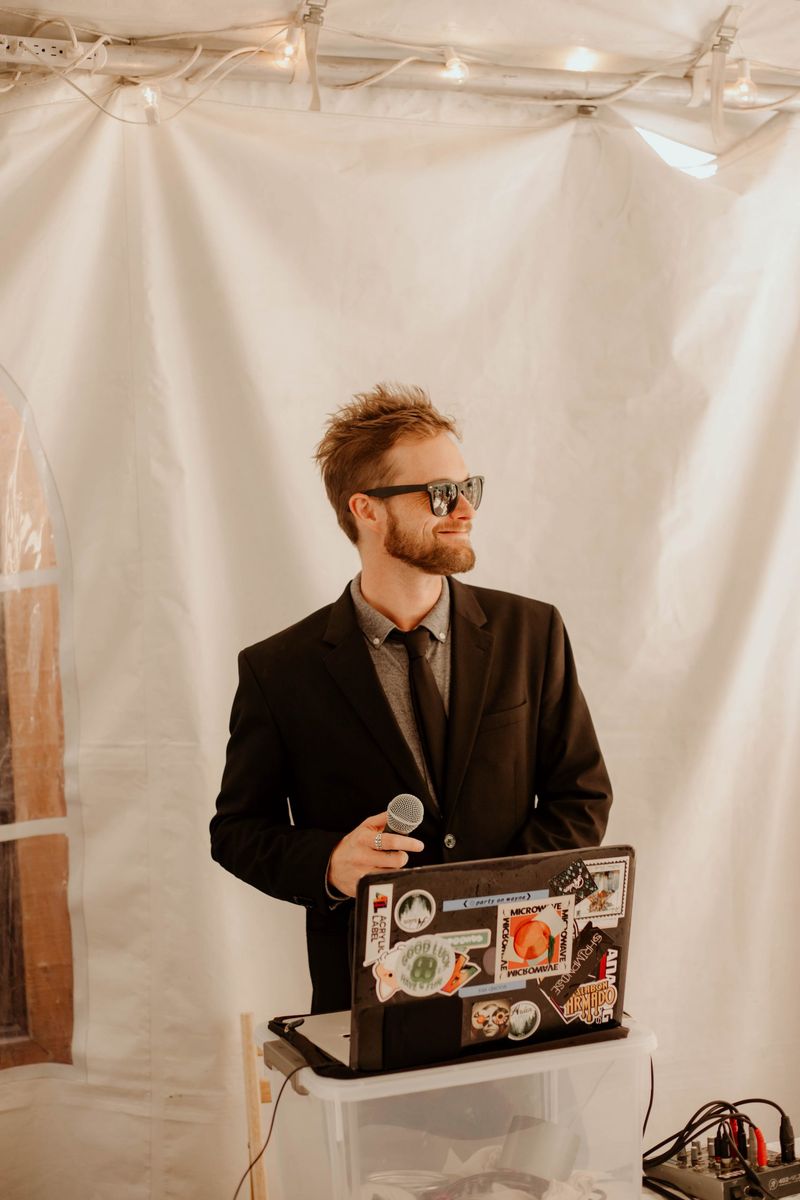Max smiling with a microphone behind his sticker-covered laptop inside a white reception tent